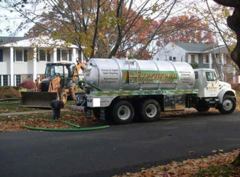 Evergreen Cesspool vacuum truck pumping a cesspool in front of a Long Island home, with a backhoe on site