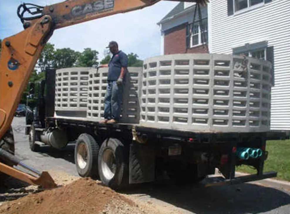 Evergreen crew member standing on a flatbed loaded with precast concrete cesspool rings beside a backhoe
